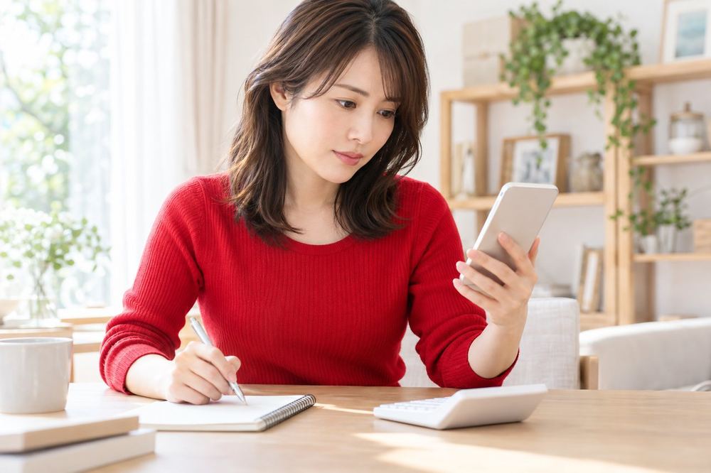 30代の女性が机で電卓とスマホの残高画面を見比べて、月額の応援のしくみをメモに整理している