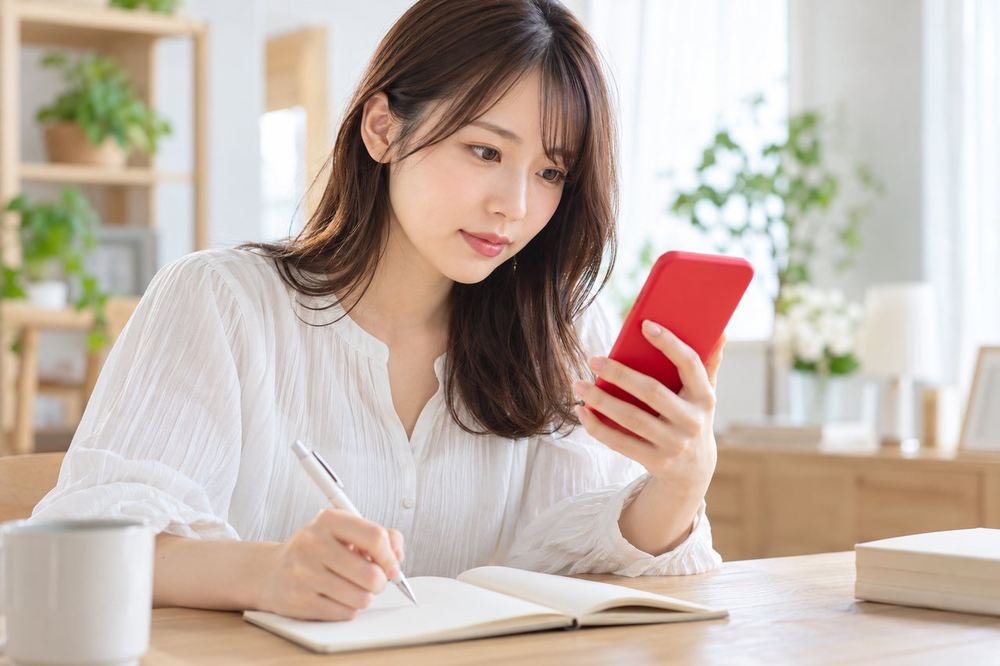 明るい部屋でスマホを見ながら、ノートに気になる点を書き出している20代の女性