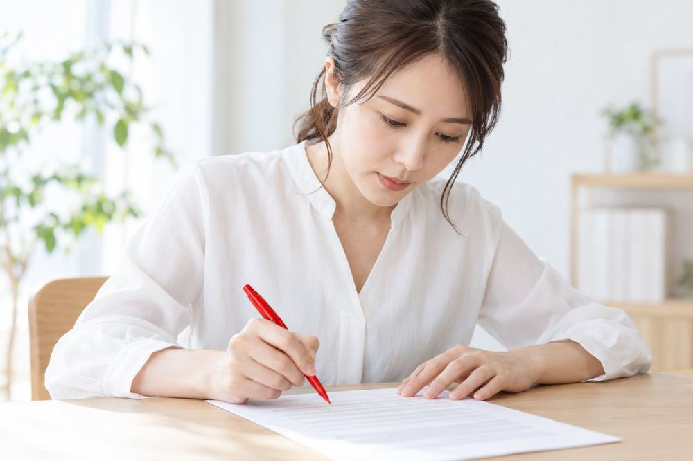 机の上で契約書のような書類を、赤いペンを片手に一行ずつ読んでいる女性