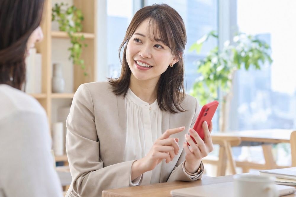 明るいオフィスの一角で、マネージャーの女性が笑顔でスマホの画面を見せながら話している