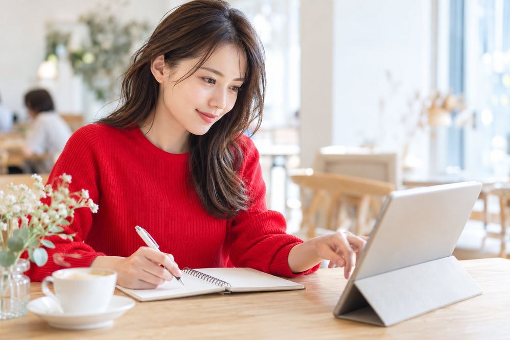 女性がカフェのテーブルでタブレットを開き、気になる項目を選びながらメモを取っている